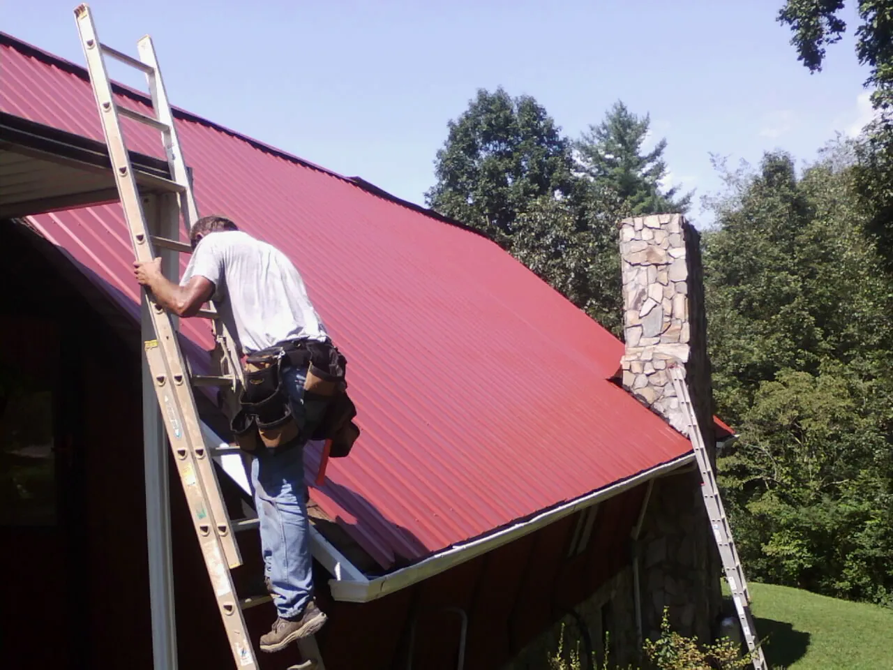 Roofer on a ladder beside a red metal roof and stone chimney — Soffit & Fascia Repair by Roofing Brothers in Beulaville
